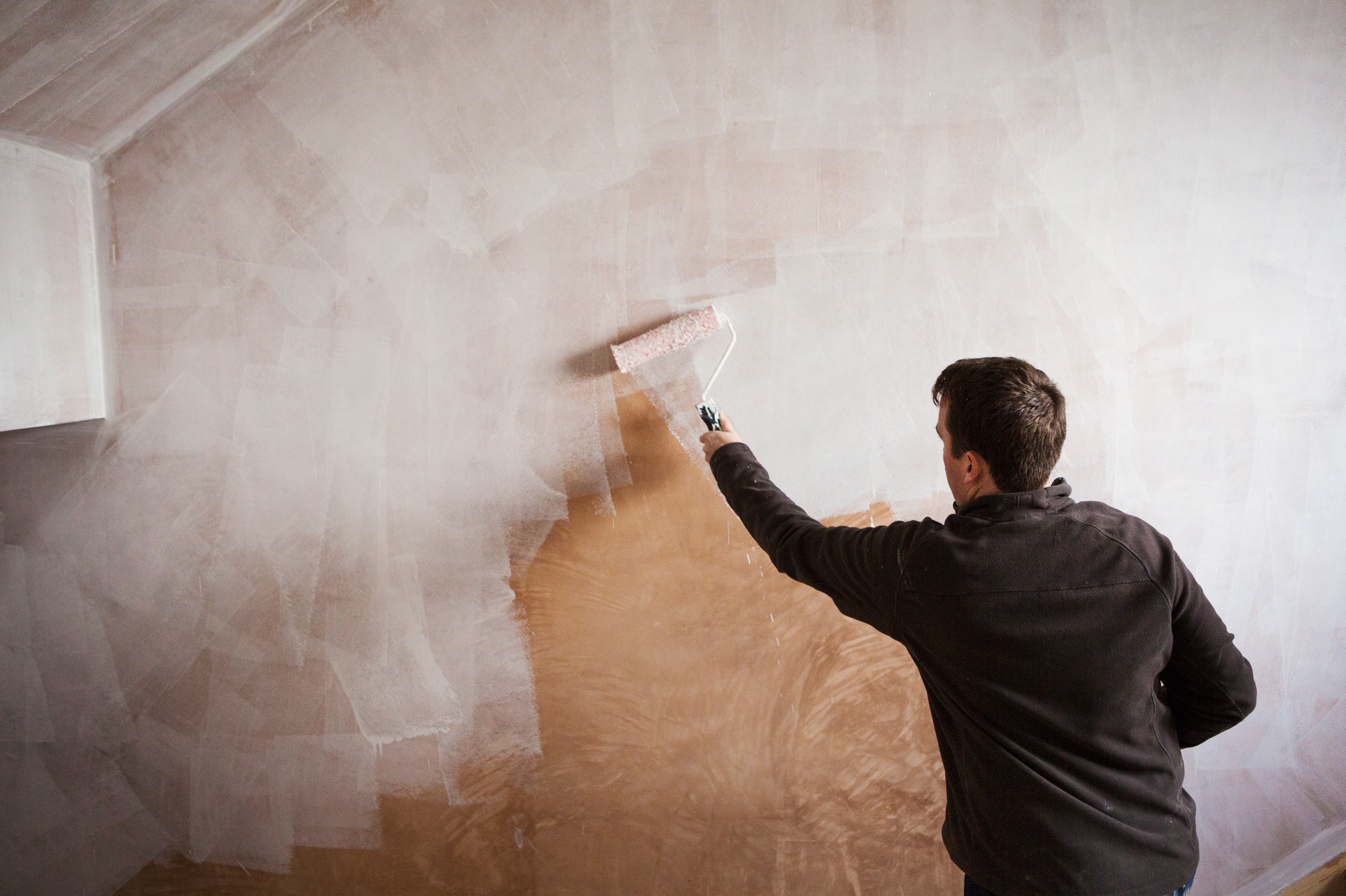 A builder, a painter holding a paint roll, painting a wall with white paint.